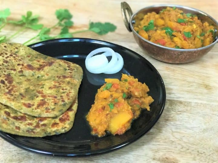 Pumpkin curry made in pressure cooker served in a black plate with two parathas and onions. 