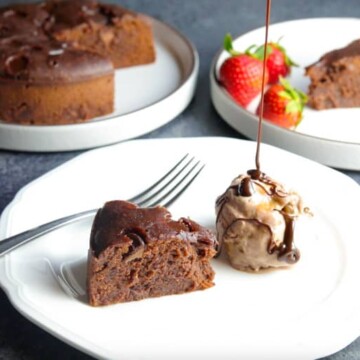 A piece of Instant Pot Brownie in a plate with ice-cream and chocolate sauce dripping, and the whole brownie cake in the back.