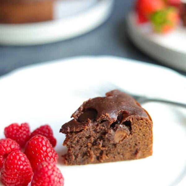 A piece of Instant Pot Brownie in a plate with raspberries, and the whole brownie cake in the back.