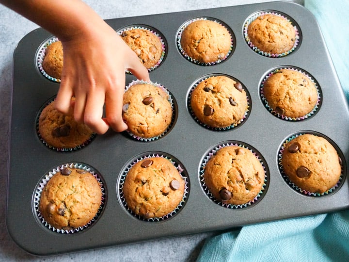 Perfectly baked Zucchini Chocolate Chip Muffins in a baking tray, one being picked by hand.