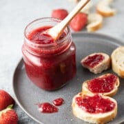 Strawberry Jam in a glass jar on a plate, along with jam spread on sliced bread.