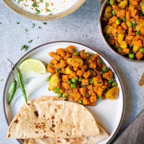 Aloo Gajar Matar (Potato, Carrots & Peas) along with roti (indian flatbread) in a white plate along with yogurt raita on the side.