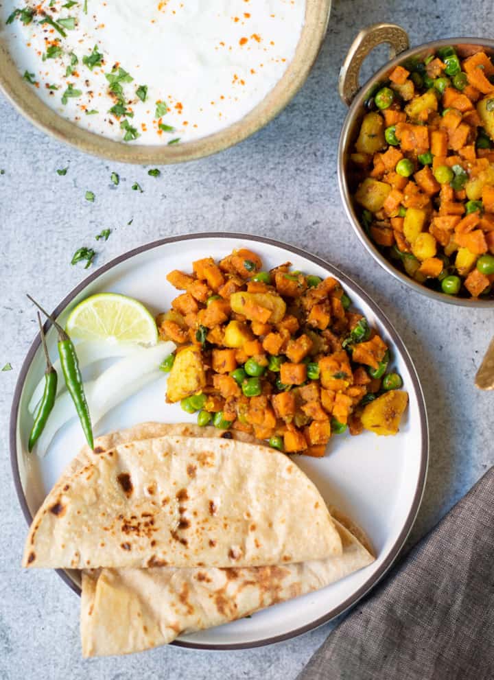 Aloo Gajar Matar (Potato, Carrots & Peas) along with roti (indian flatbread) in a white plate along with yogurt raita on the side. 