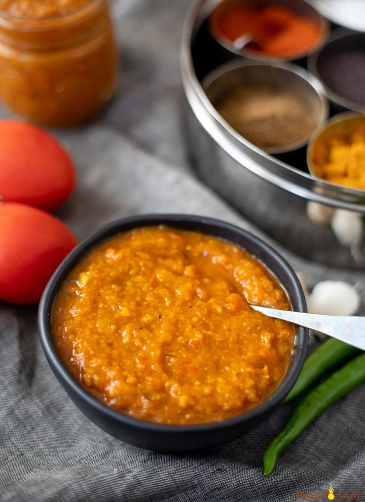 Indian curry sauce made with onions and tomatoes in a black bowl with spices and tomatoes in the back