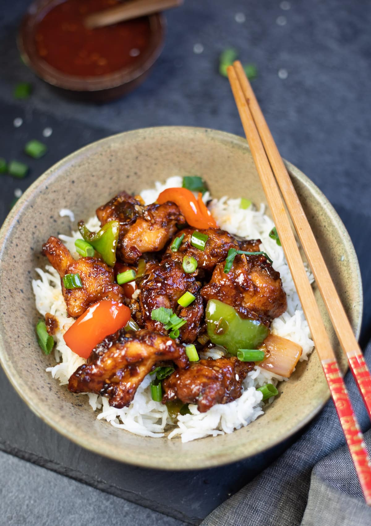 Chilli Chicken over white rice in a bowl, with spicy chili sauce in a small bowl