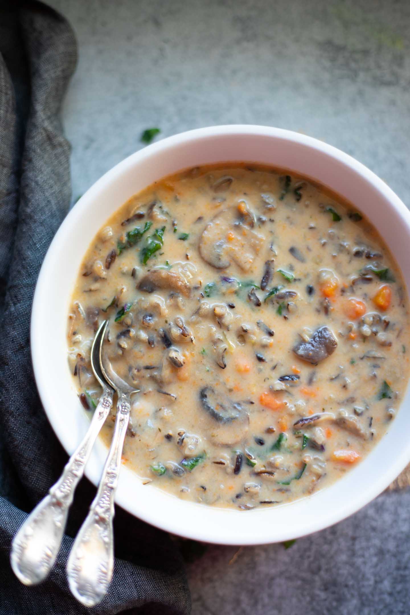 a creamy brown soup with rice and vegetables in a white dish with two silver spoons