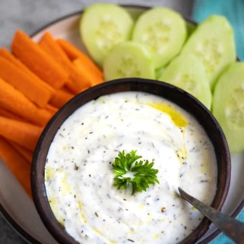 yogurt dip garnishes with parsley in a bowl along with carrots and cucumbers on the side to dip