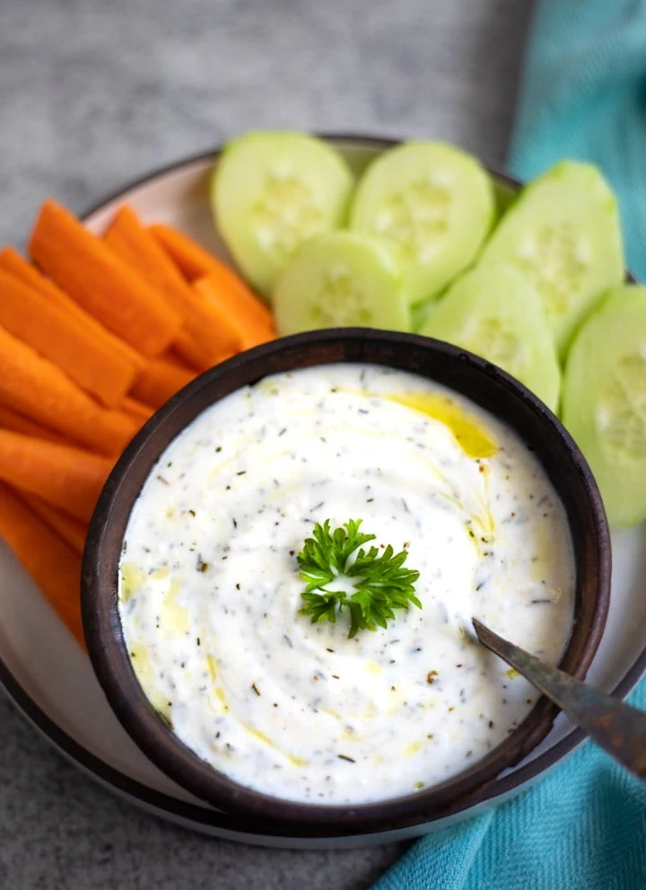 yogurt dip garnishes with parsley in a bowl along with carrots and cucumbers on the side to dip