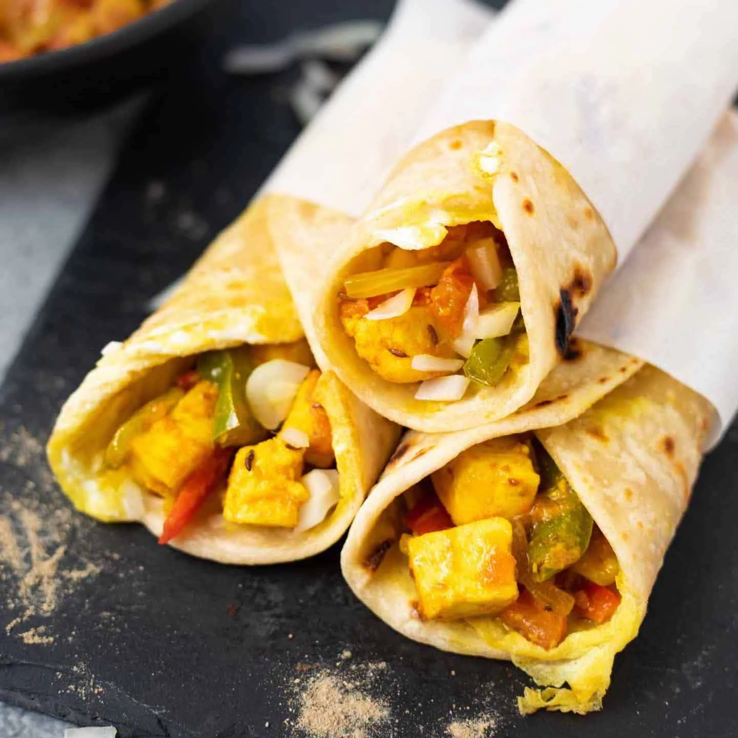 Three paneer Kathi roll wraps on a black base with a bowl of the filling in the back
