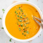 Creamy Carrot Soup in a white bowl garnishes with cilantro, red pepper flakes and sesame seeds