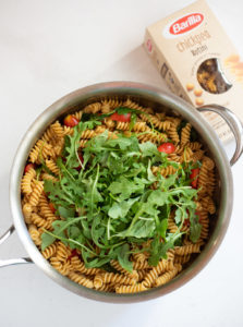 Rotini with cherry tomatoes and topped with baby arugula leaves in a large pan with barilla chickpea pasta box on the side