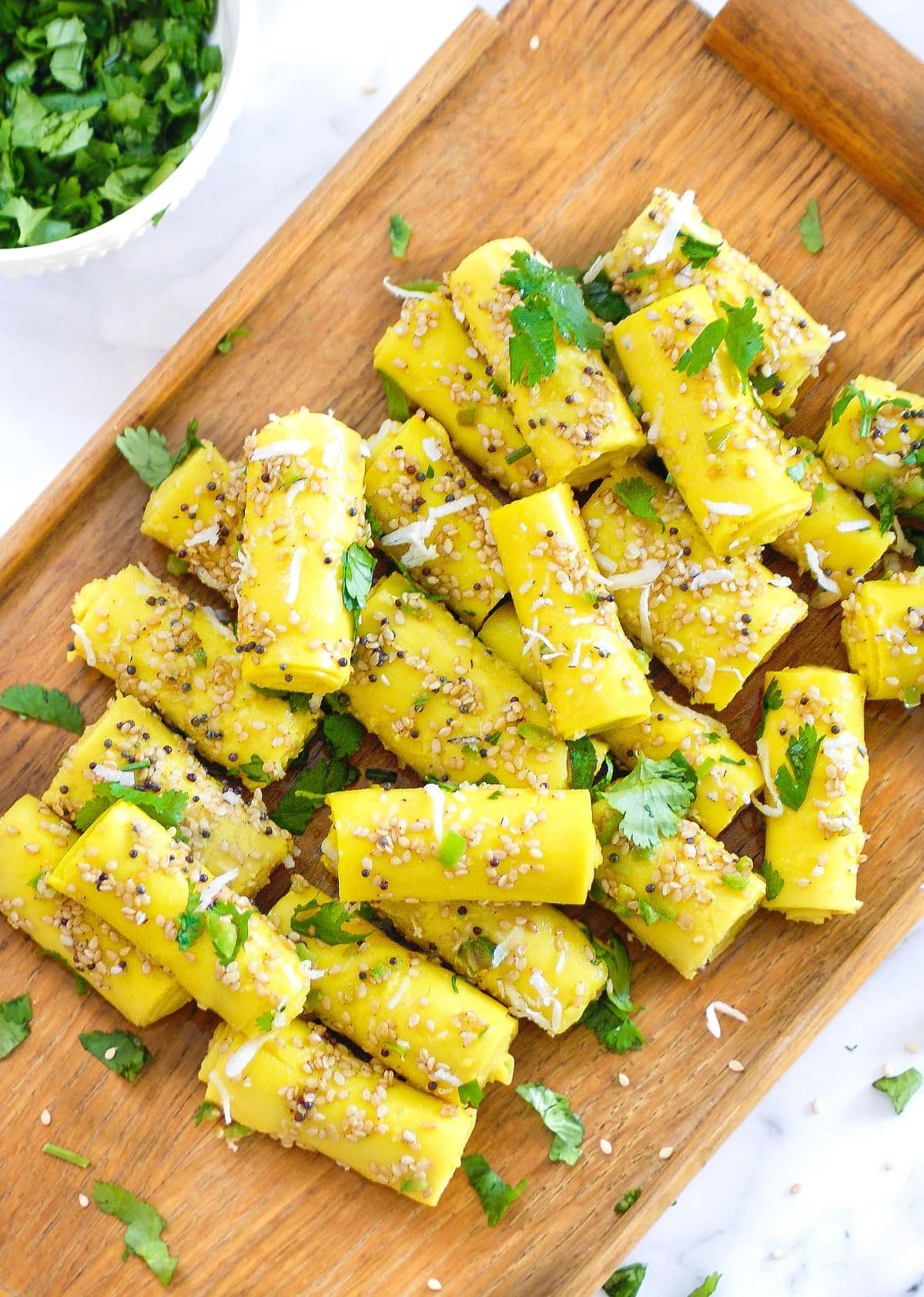 Khandvi on a wooden platter topped with a tempering of sesame seeds and garnished with cilantro