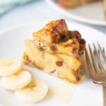 A piece of banana bread pudding drizzled with syrup and banana slices on the side.