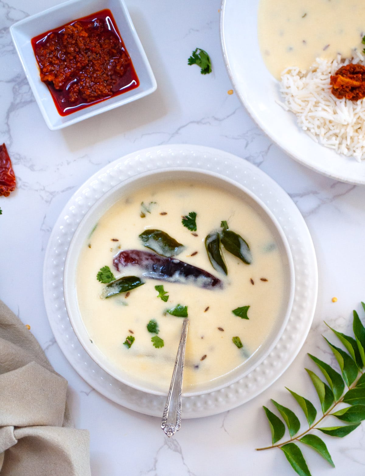 Gujarati kadhi served in a white bowl garnished with cilantro. Pickle and rice served on the side. 