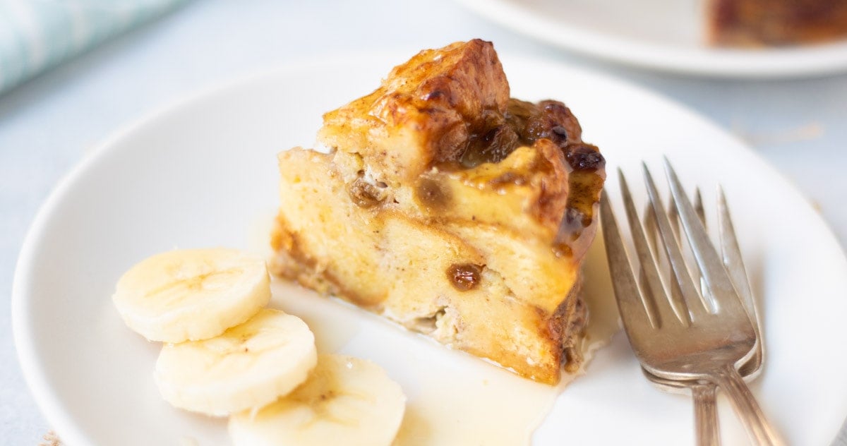 A piece of banana bread pudding drizzled with syrup and banana slices on the side.