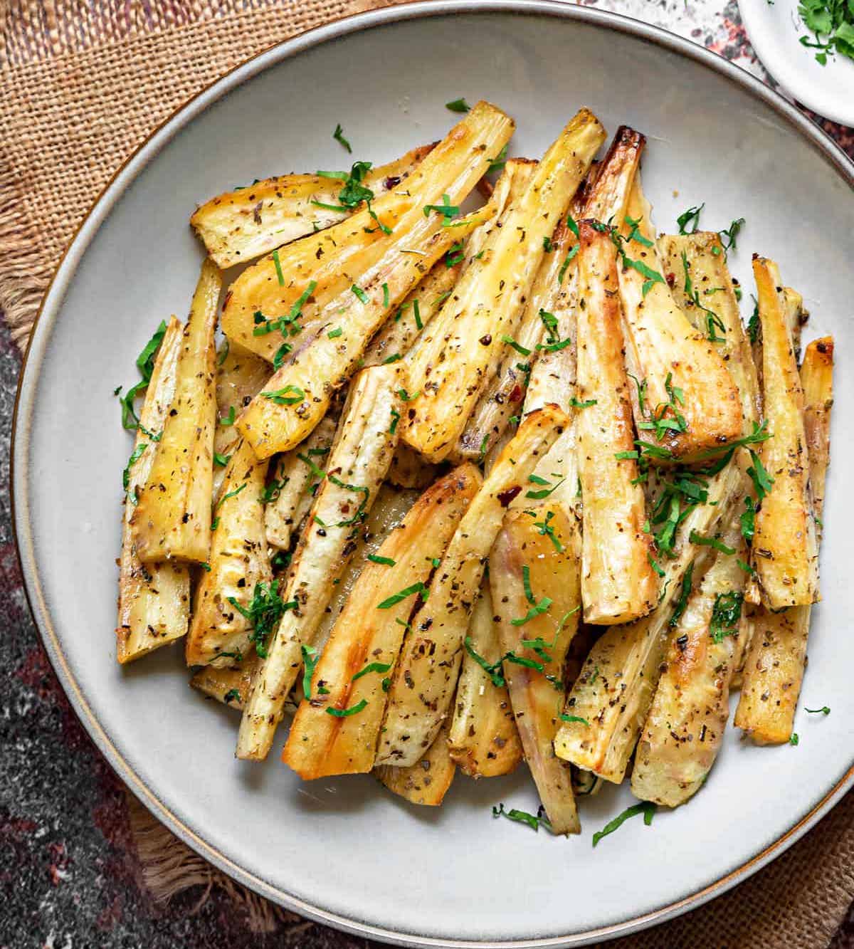 overhead shot of a pile of roasted parsnip batons on a white plate and sprinkled with fresh parsley