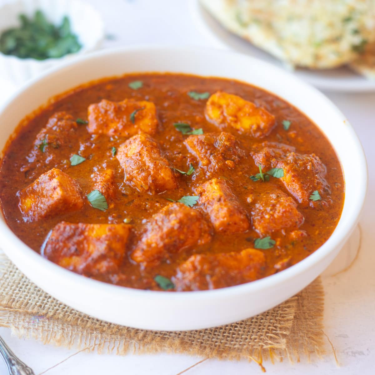 Paneer vindaloo curry in a white bowl with naan in the back.