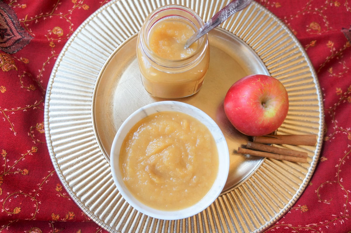 top view of Applesauce in a white bowl and in a glass jar with apple and cinnamon on side in a silver plate