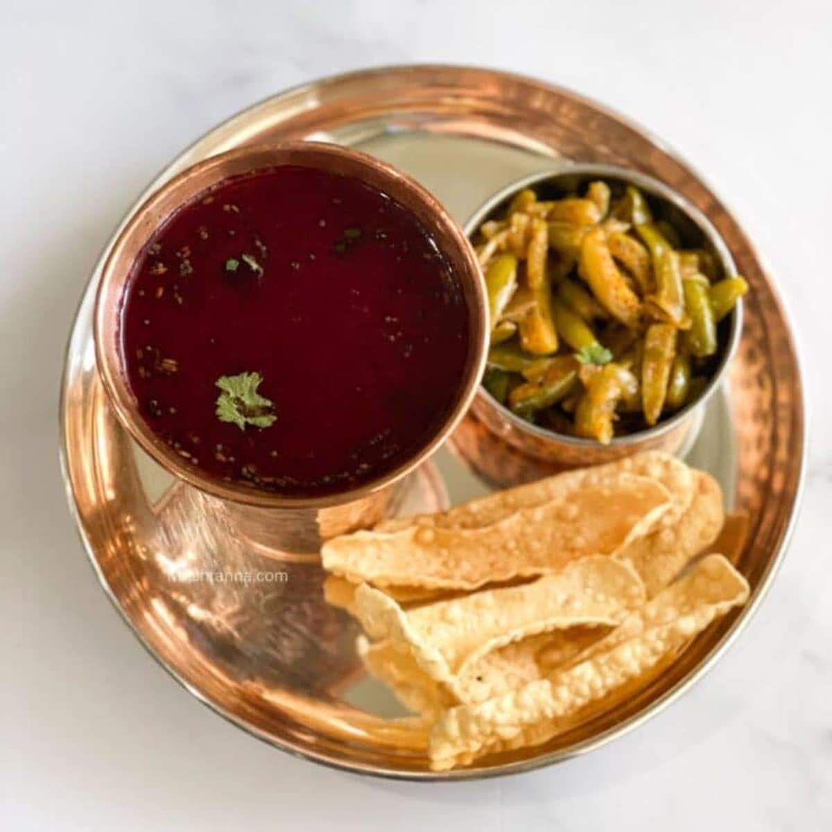 A copper glass is filled with beetroot rasam and placed on the silver plate with stir fry.