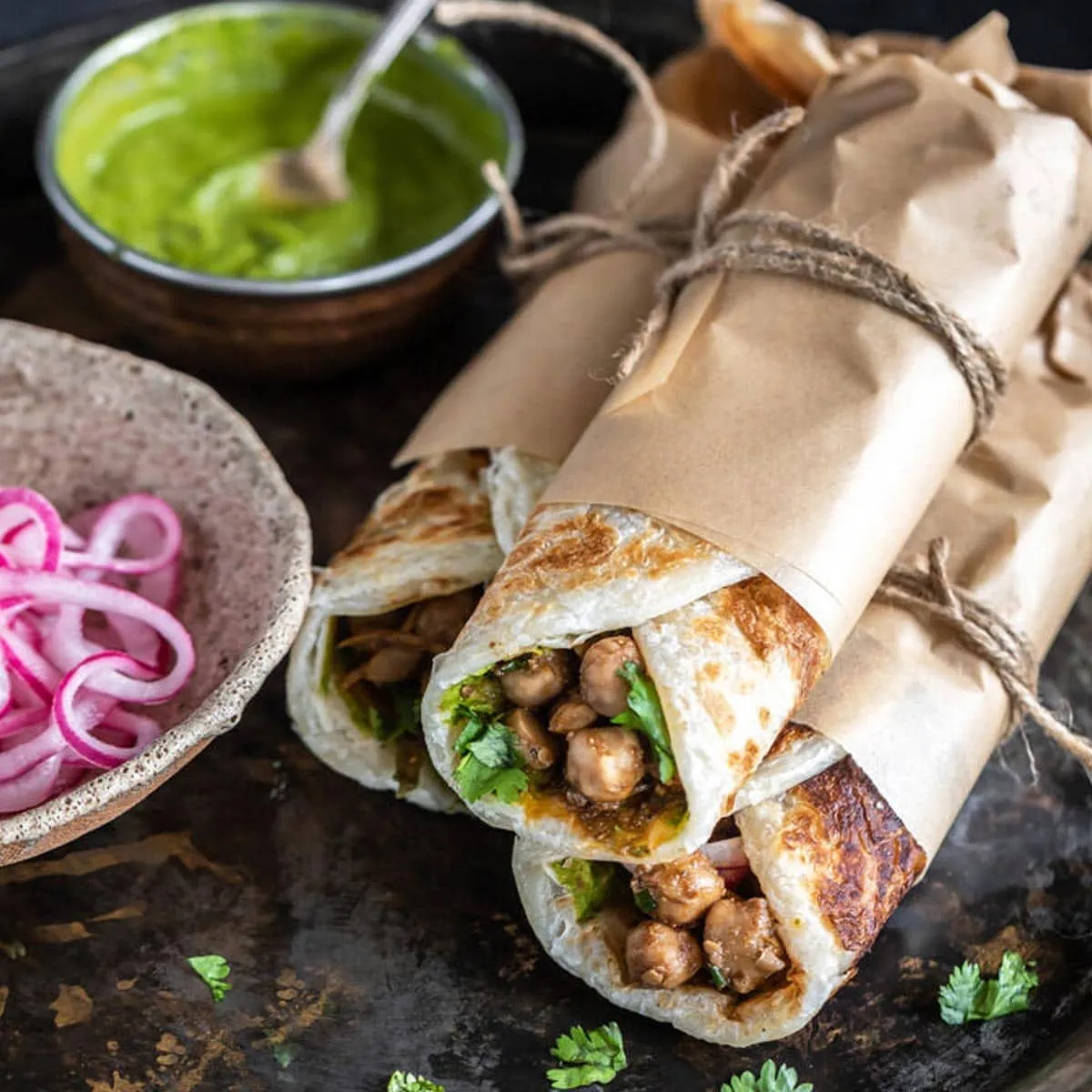 three wraps rolled in parchment paper and tied with thread stacked together with a bowl of chutney and pickled onions on the side