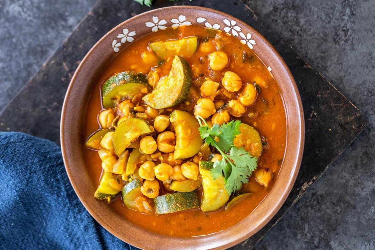 zucchini and chickpea curry served in a brow color bowl and a blue napkin placed in the background