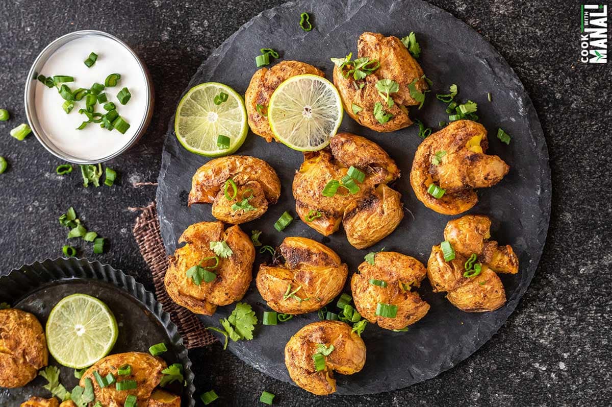 masala smashed potatoes served on a circular cheese board with lime wedges placed on the side. There's also a white color dip in a bowl in the background