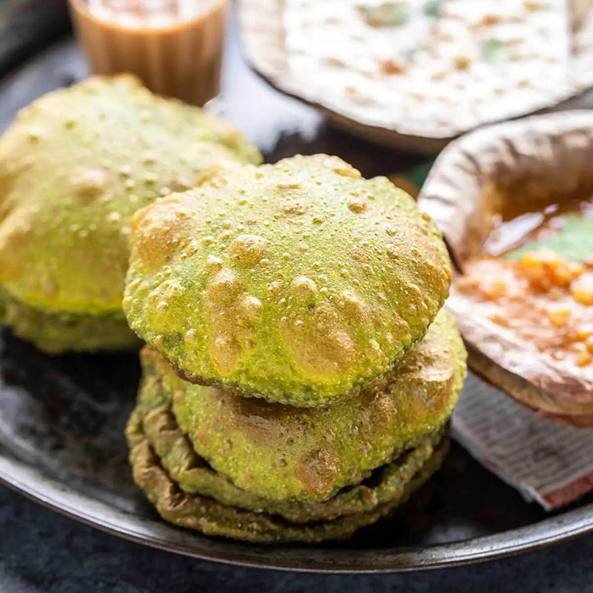 stack of puris placed in a plate along with glass of chai and bowls of curry and raita