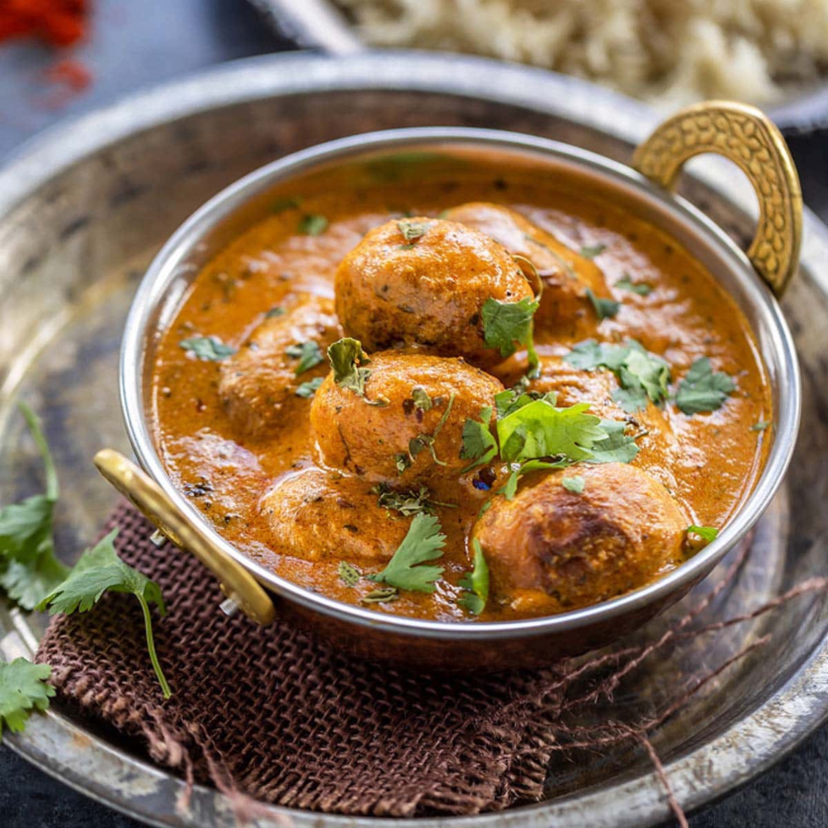 dum aloo served in a copper kadai garnished with cilantro with a plate of rice in the background