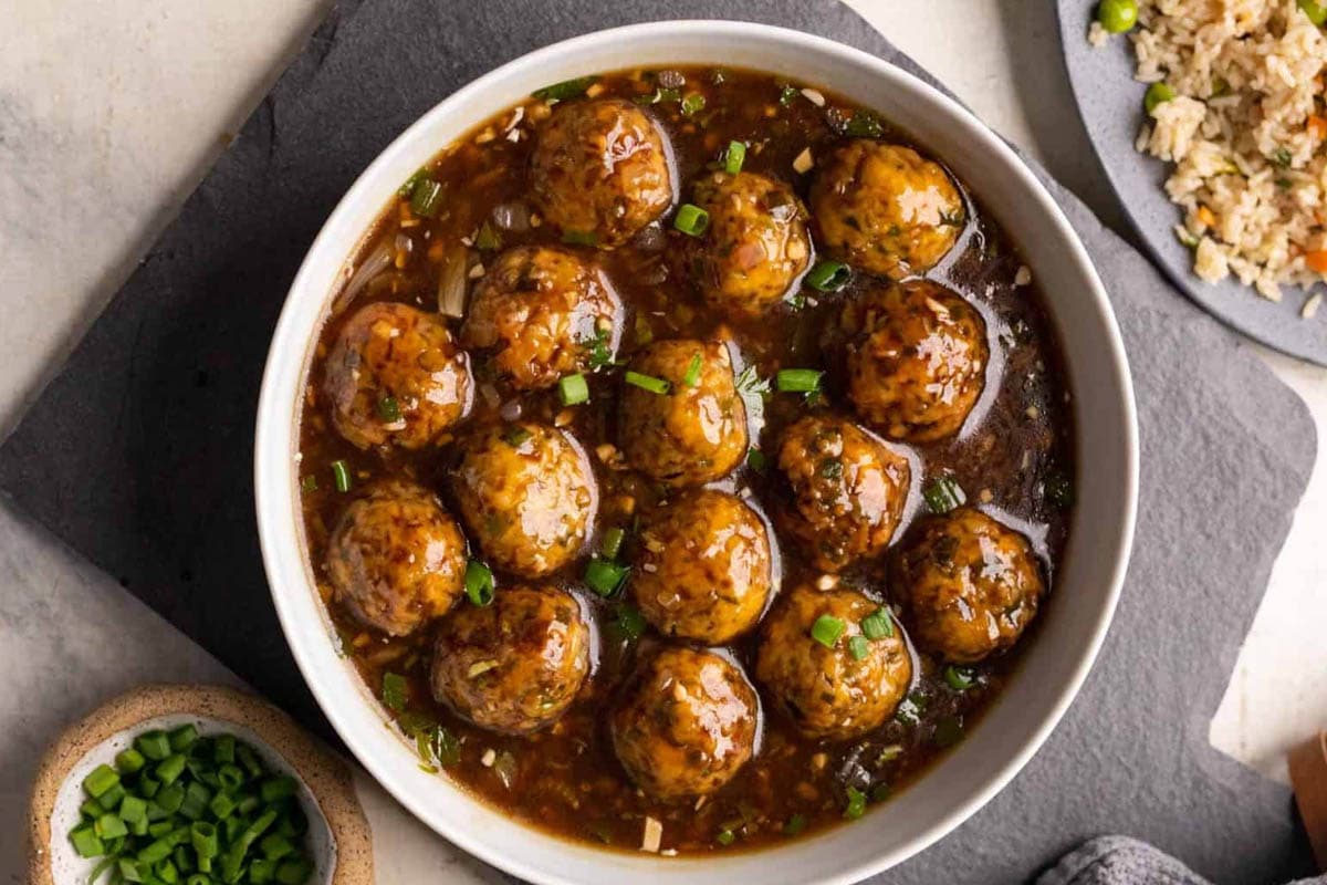 Veg manchurian served in a white bowl with fried rice and spring onions on the side