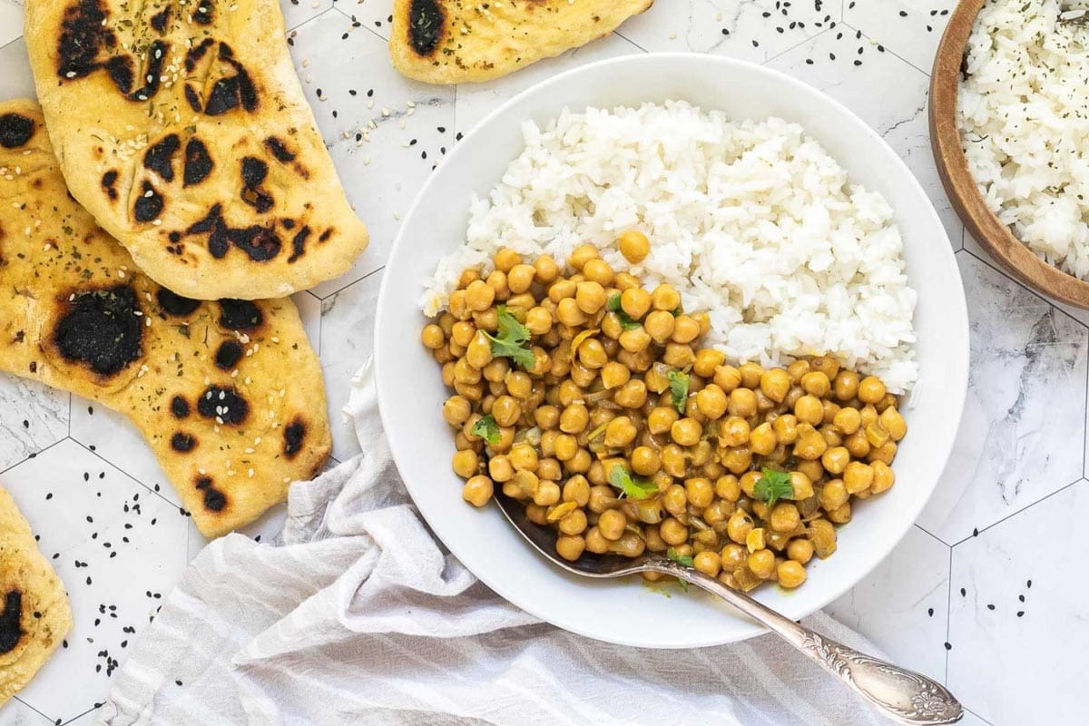 A white plate with white rice and chickpeas in a creamy orange sauce sprinkled freshly chopped cilantro. A spoon is placed inside. Naan bread and a bowl of rice are next to it.