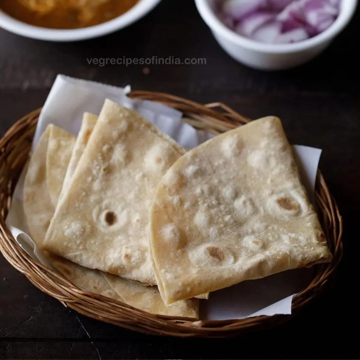 rumali roti folded and served in a basket with a bowl of curry and a bowl of onions kept in the background. 