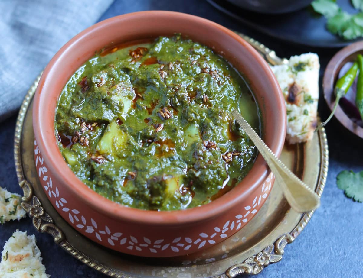 Saag Aloo made in the instant pot served in a earthen bowl with the tempering in the back and naan on the side.