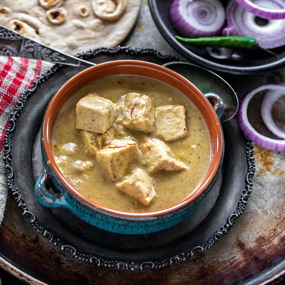 creamy shahi tofu curry served in a blue bowl with sliced red onion rings on one side and tandoori roti placed in the background