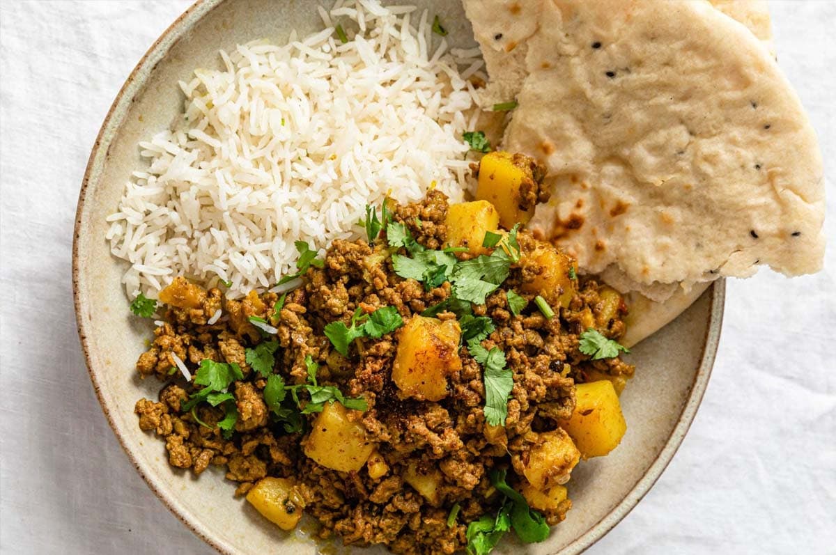 Aloo Keema (Ground Beef and Potato Curry) in a beige bowl served with naan and rice and garnished with cilantro