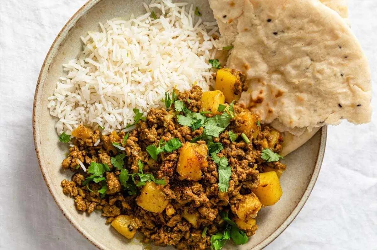 Aloo Keema (Ground Beef and Potato Curry) in a beige bowl served with naan and rice and garnished with cilantro
