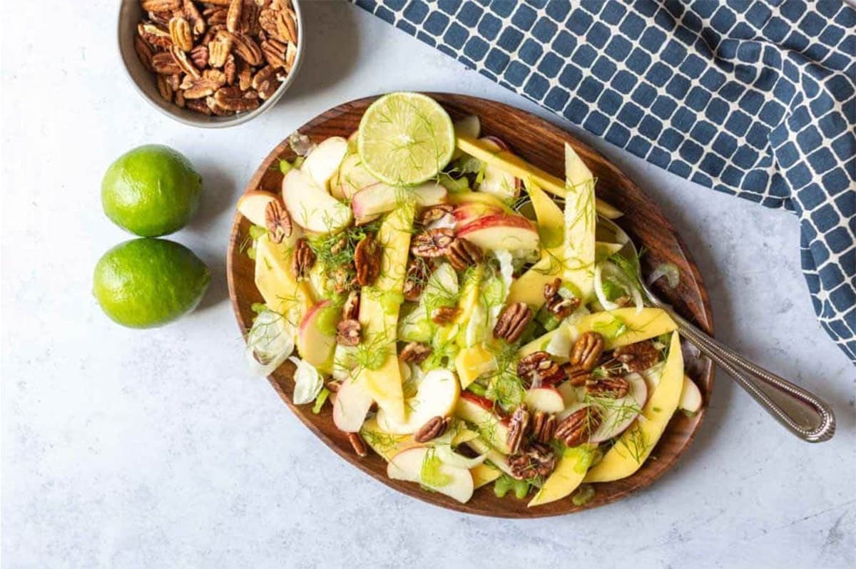 mango apple and fennel salad on a brown plate with bowl of pecans on side