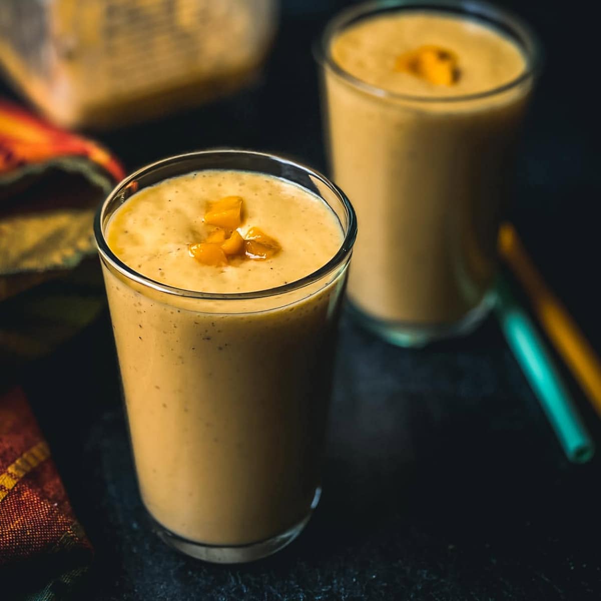 Two glasses filled with Mango Banana Smoothie with green and orange straws on the side