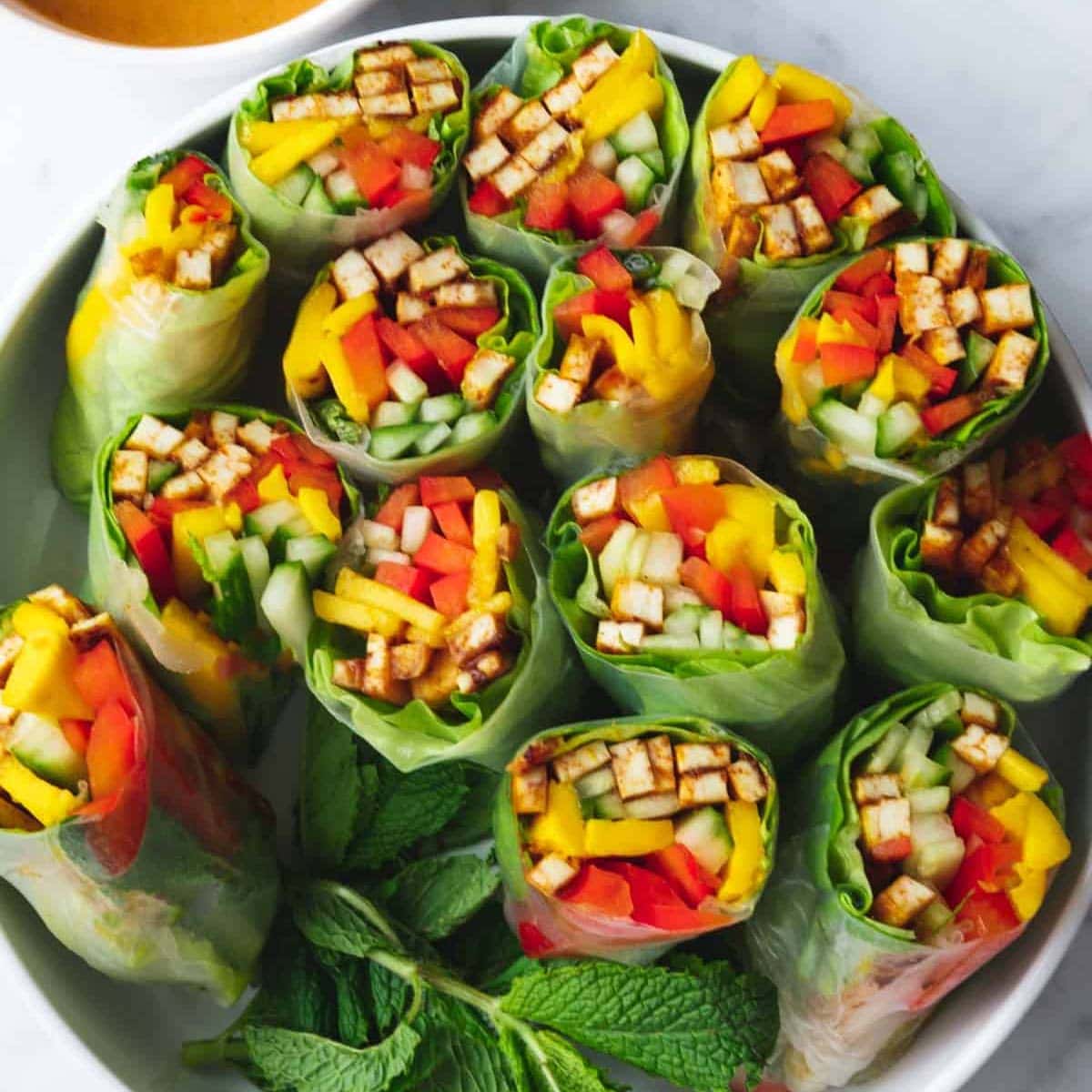 7 summer rolls cut in half in a white bowl. Filled with tofu, mango, cucumber, red bell pepper, lettuce, and mint. Served with peanut dipping sauce.