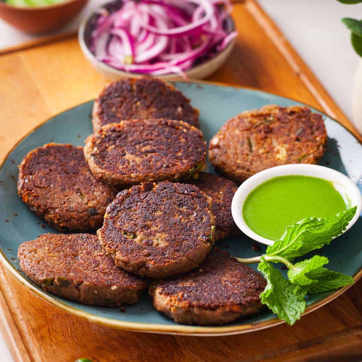 Shami Kebab on a blue plate served with green chutney, cucumber and pickled onions.