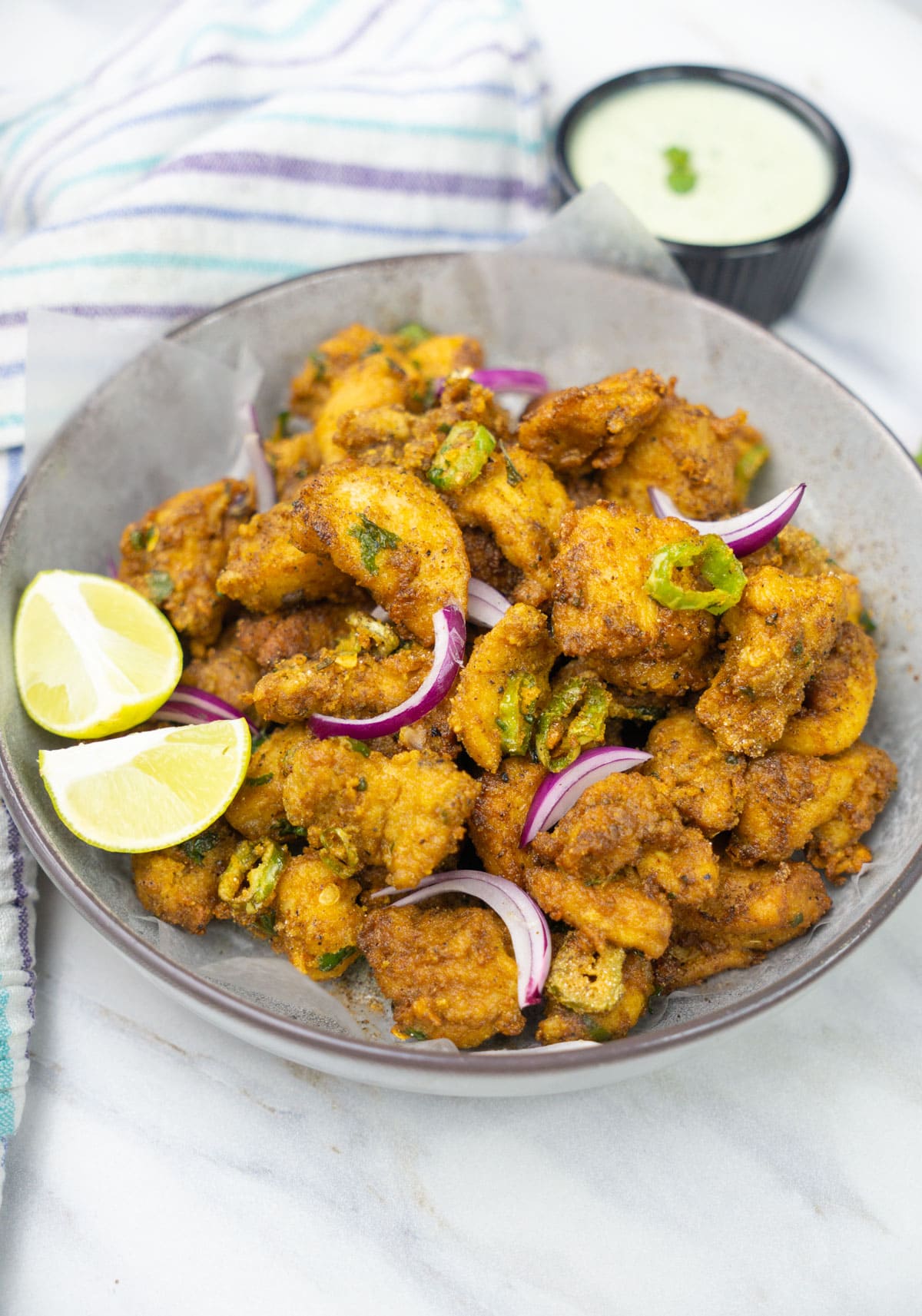 Crispy chicken pakora in a bowl topped with sliced onions and lime wedges. 