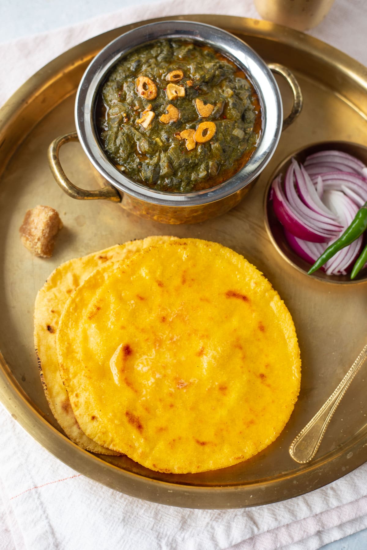 Makki ki Roti served with Sarson ka saag in a brass plate with onion, green chili and jaggery on the side. 