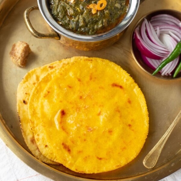 Makki ki Roti served with Sarson ka saag in a brass plate with onion, green chili and jaggery on the side.