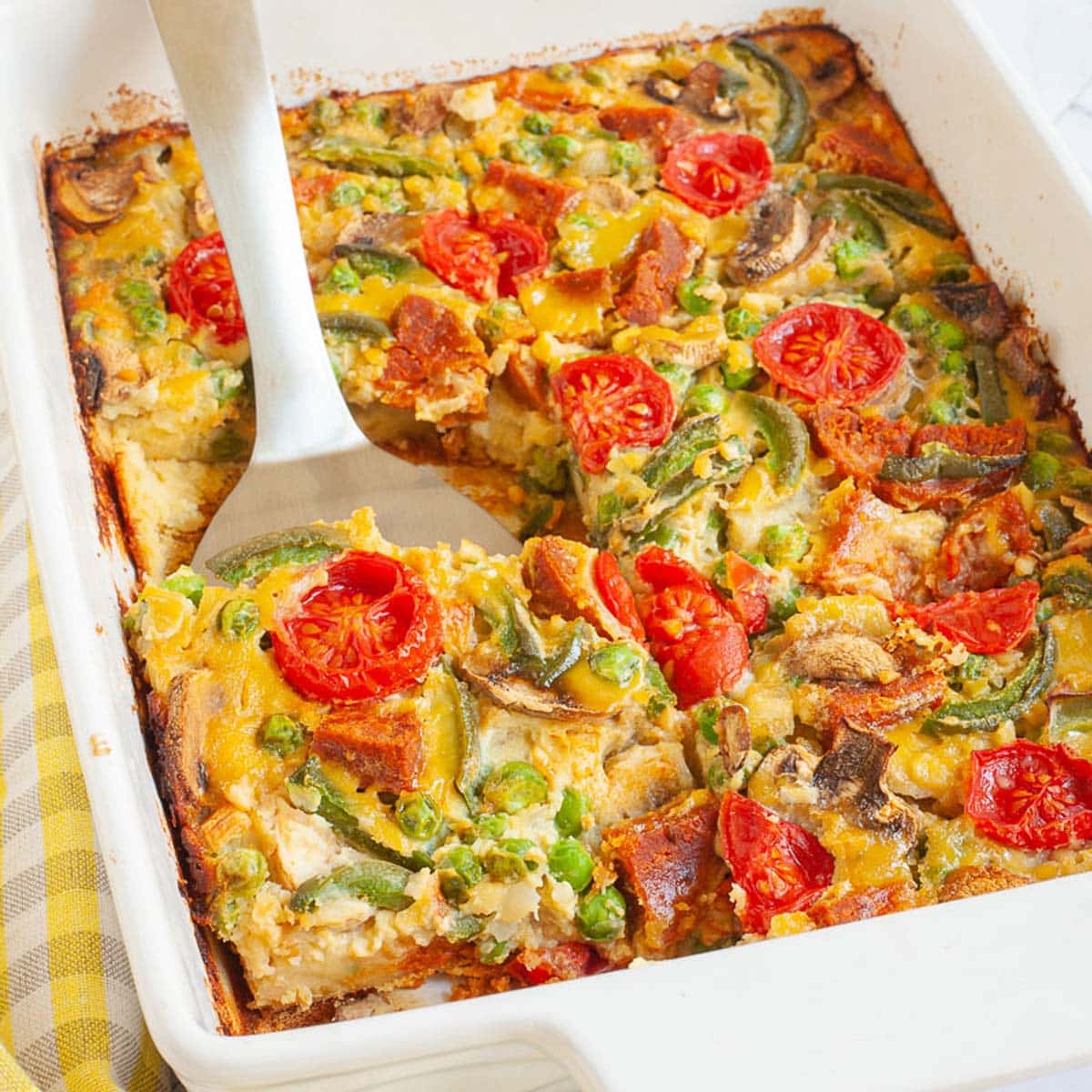 Cherry tomatoes, mushroom slices, bell pepper slices on top of a yellow cake-like dough is in a white casserole dish. A slice served on a white plate is in the background