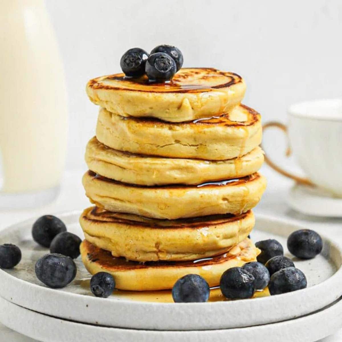 Stack of 6 thick chickpea flour pancakes topped with blueberries and maple syrup on a small plate. A jug of milk and a coffee cup are in the background.