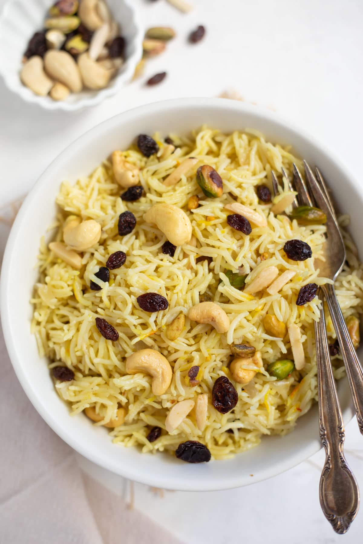 Rice pilaf with nuts and raisins in a white serving bowl, garnished with roasted dry fruits and bowl of dry fruits on the side