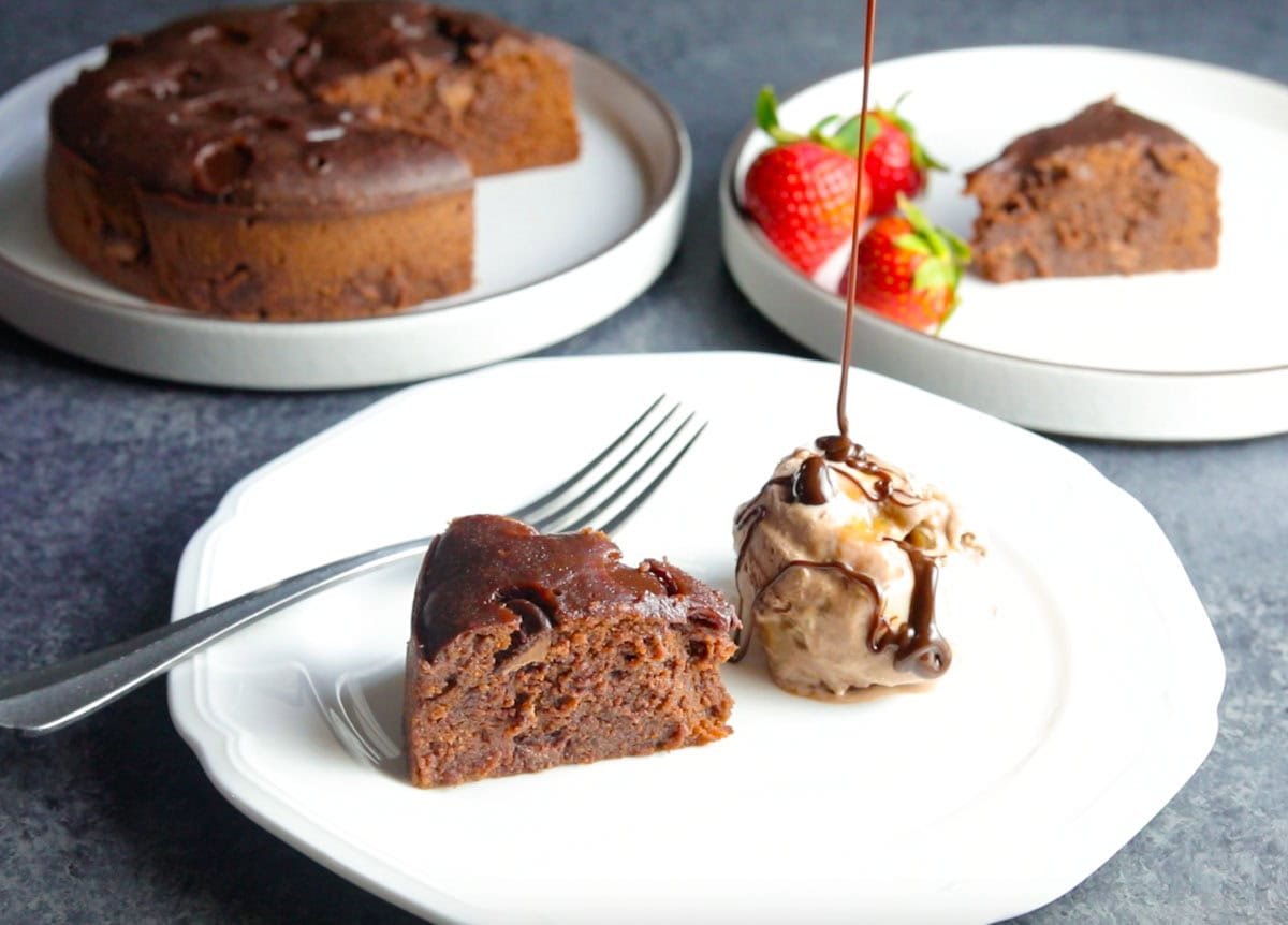 A piece of Instant Pot Brownie in a plate with ice-cream and chocolate sauce dripping, and the whole brownie cake in the back and fresh strawberries.