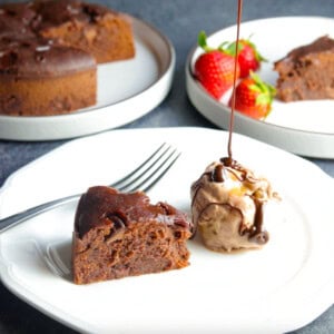 A piece of Instant Pot Brownie in a plate with ice-cream and chocolate sauce dripping, and the whole brownie cake in the back.