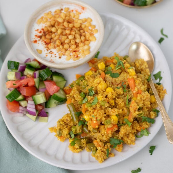 quinoa pilaf with vegetables, cucumber tomato salad, and raita on a white plate