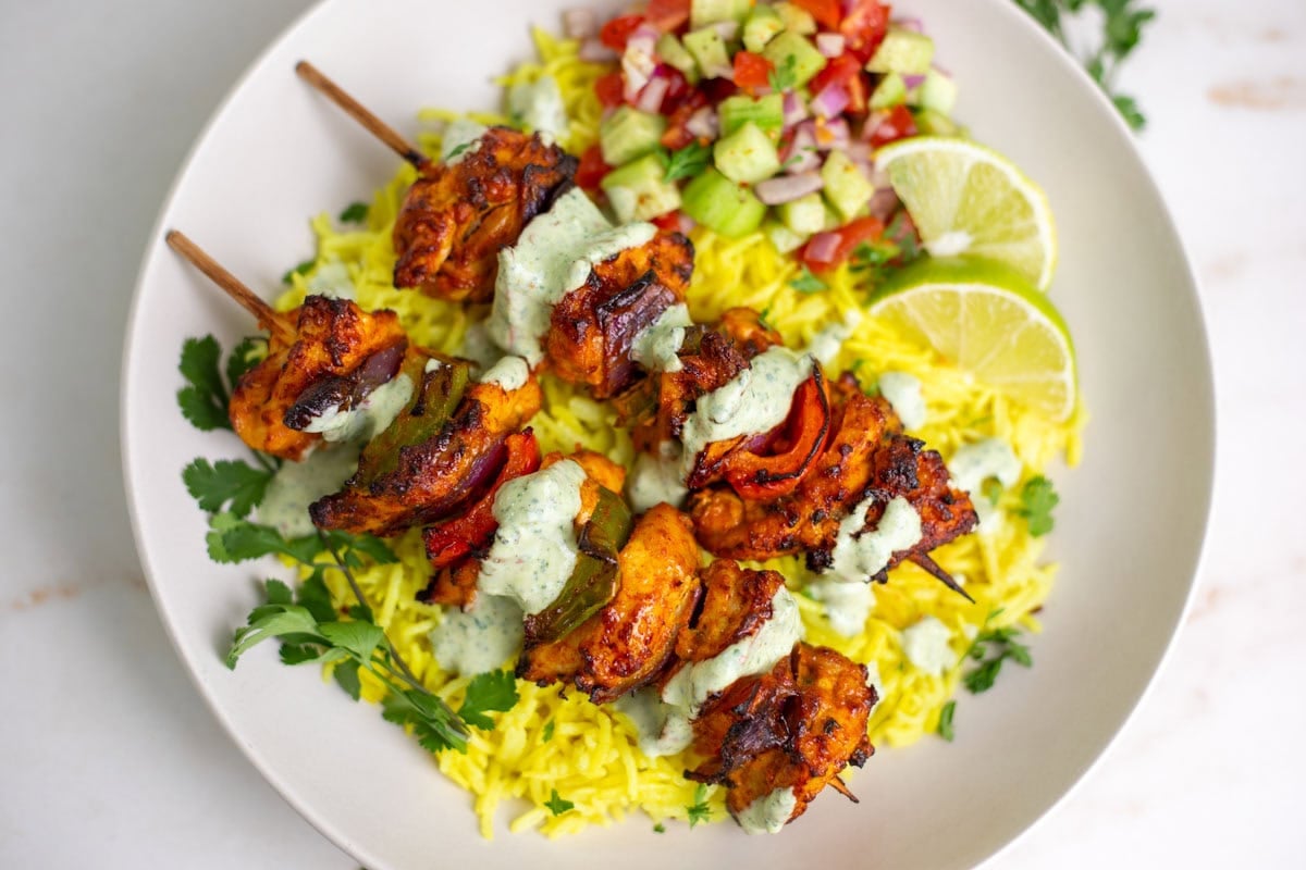 Chicken Tikka Kebab Skewers drizzled with chutney served over yellow rice with a side of kachumber salad.