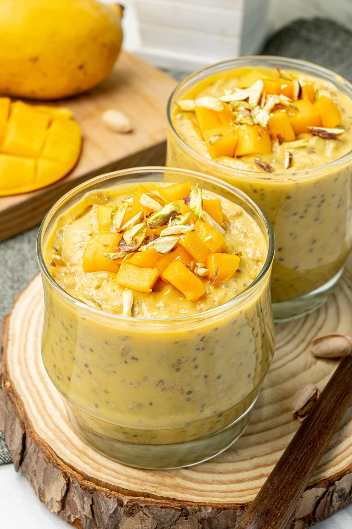 Creamy mango overnight oats served in two cups, topped with fresh mango cubes and slivered pistachios.